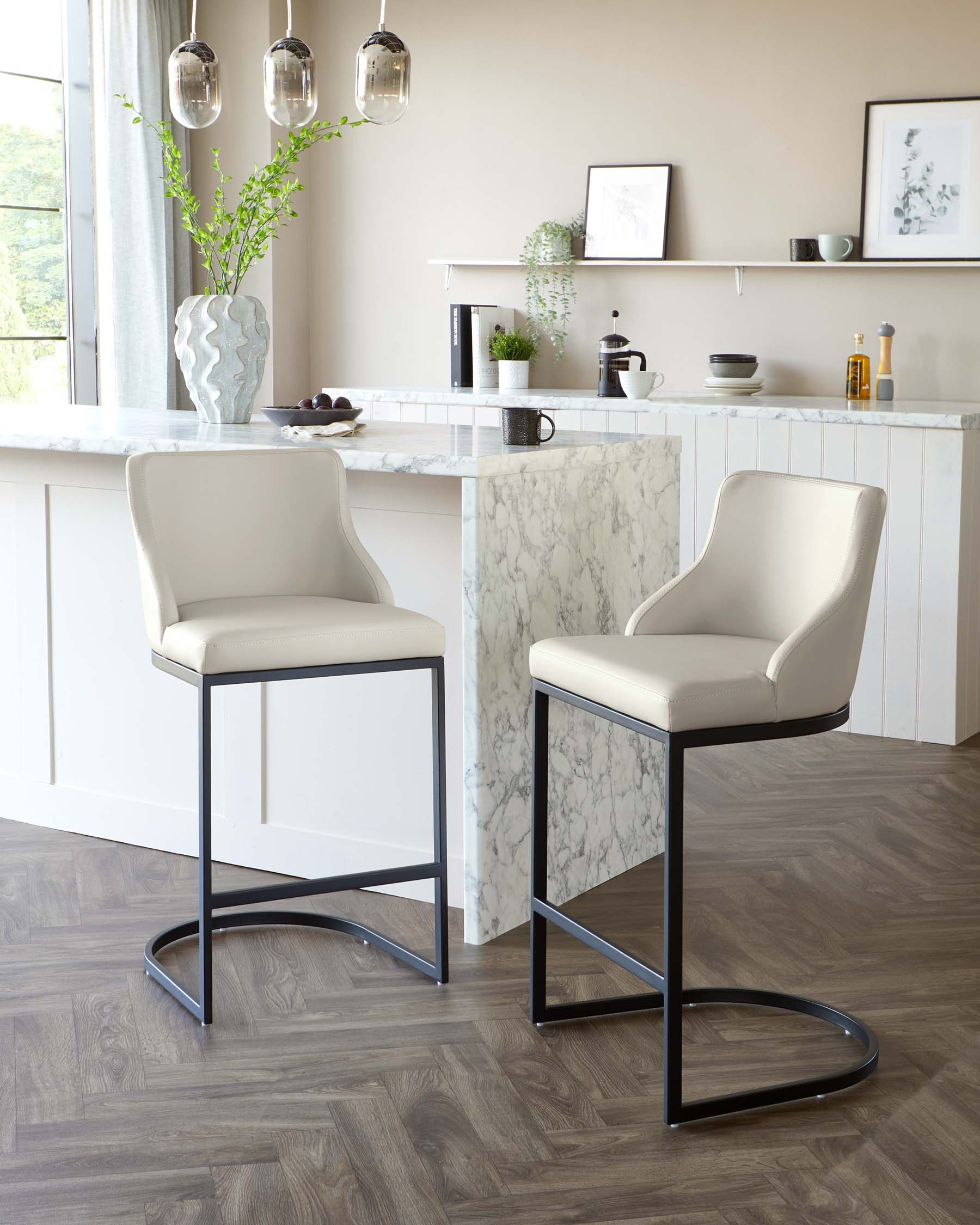 Two modern bar stools with sleek cream upholstery and black metal frames next to a kitchen island with a marble countertop.