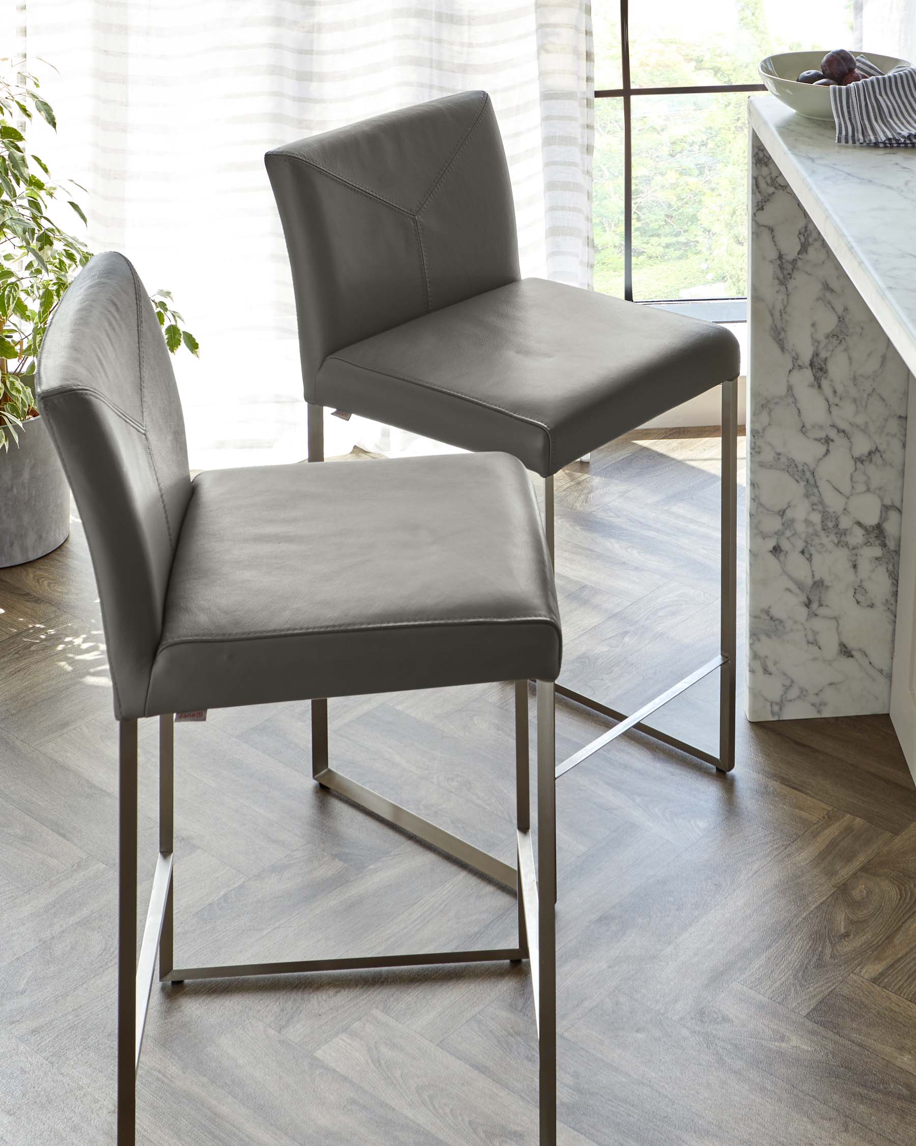 Two modern grey leather bar stools with sleek metal legs positioned near a marble countertop in a bright room with wooden flooring.