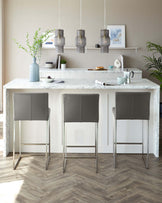 Three contemporary bar stools with grey upholstery and slender metallic legs in front of a marble top kitchen island.