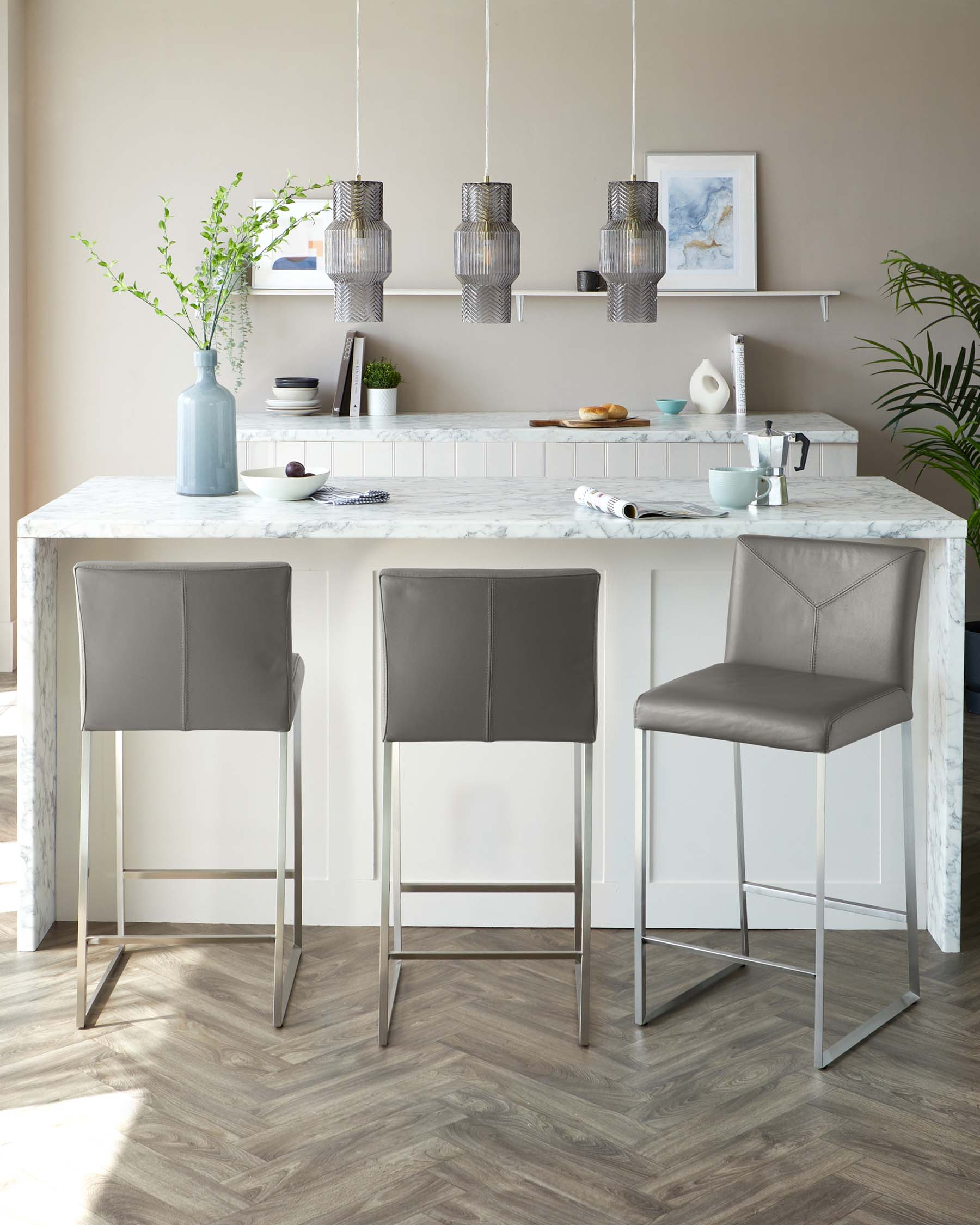 Modern kitchen interior featuring three sleek bar stools with grey upholstery and slender, metallic legs, arranged neatly at the kitchen island with a white marble top.