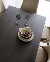 Modern minimalist dining table with a dark wood finish and simple, clean lines, accompanied by elegant beige upholstered dining chairs with a smooth, curved backrest. The table is set with two smoky wine glasses and a refined plate setting featuring grapes.