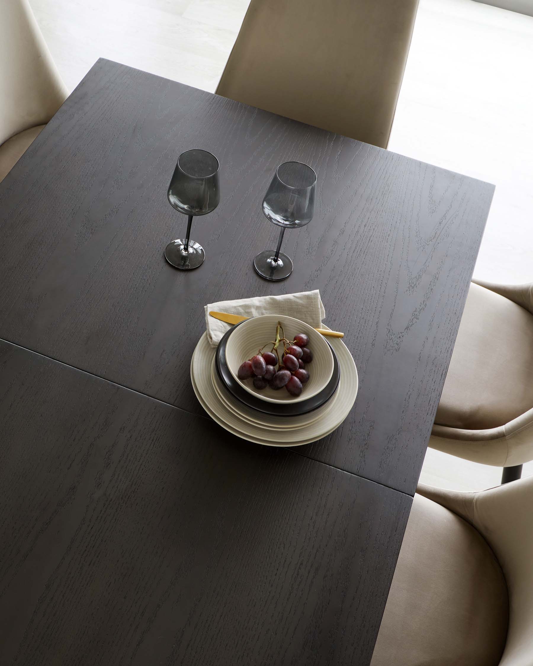 Modern minimalist dining table with a dark wood finish and simple, clean lines, accompanied by elegant beige upholstered dining chairs with a smooth, curved backrest. The table is set with two smoky wine glasses and a refined plate setting featuring grapes.