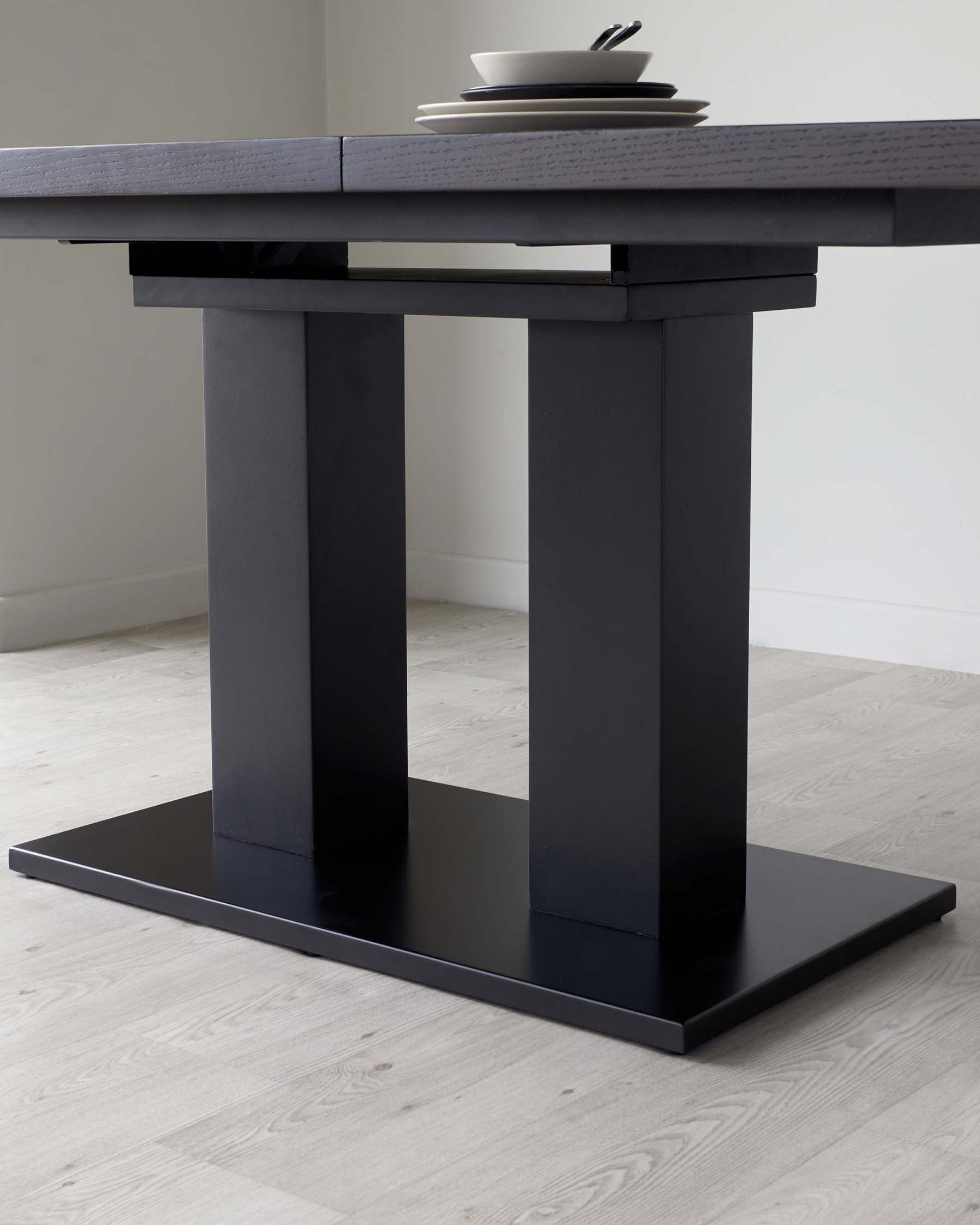 Modern minimalist black dining table with a thick rectangular top and a unique pedestal base, featuring a solid rectangular block resting on a flat horizontal platform. A set of simple white dishes with utensils rests on top of the table, highlighting its sleek and contemporary design. The table is set against a light wooden floor and a neutral wall, emphasizing its bold, monochromatic aesthetic.