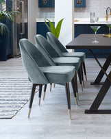 A set of four luxurious modern dining chairs with soft grey velvet upholstery and elegant dark wood legs accented with metallic tips, arranged around a contemporary dark wooden table with a black X-shaped base, set on a patterned area rug in a stylish kitchen with blue cabinetry and wooden flooring.