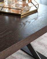 A close-up of a contemporary dark wood coffee table with a pronounced wood grain texture and a matte finish, featuring a sturdy rectangular top and an angular, minimalist metallic base with a black finish, accentuated by a gold-tone metal tray holding clear glass decor on the surface.