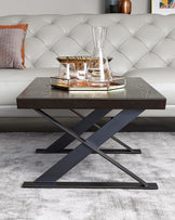 Modern rectangular coffee table with dark wood finish and distinctive X-shaped metal legs in a black matte finish, staged on a light grey area rug in front of a tufted white leather sofa with a decorative pillow. The table is adorned with a glass decanter, matching glasses on a tray, and a book.