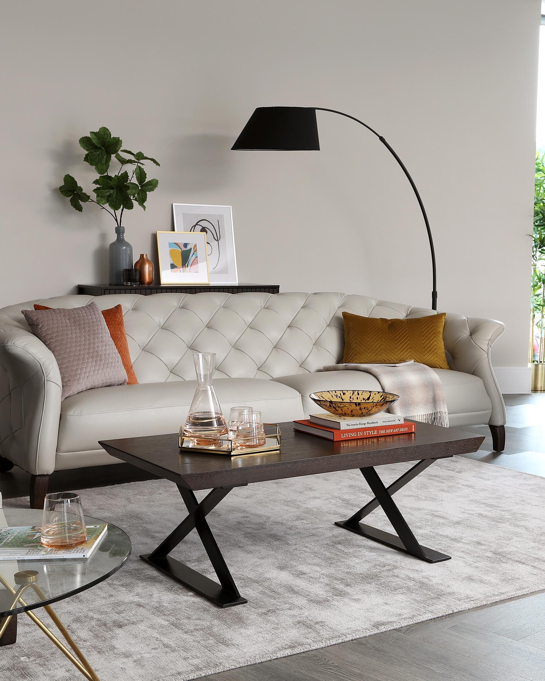 An elegant contemporary living room with a tufted, cream-colored leather sofa adorned with orange and beige throw pillows. In front of the sofa is a rectangular wooden coffee table with a unique X-shaped metal base, holding decorative items and books. A second round glass-top side table with a gold frame sits to the left. The furniture rests on a light grey area rug that complements the neutral-toned walls. An arched floor lamp with a black shade extends over the sofa to complete the stylish setup.