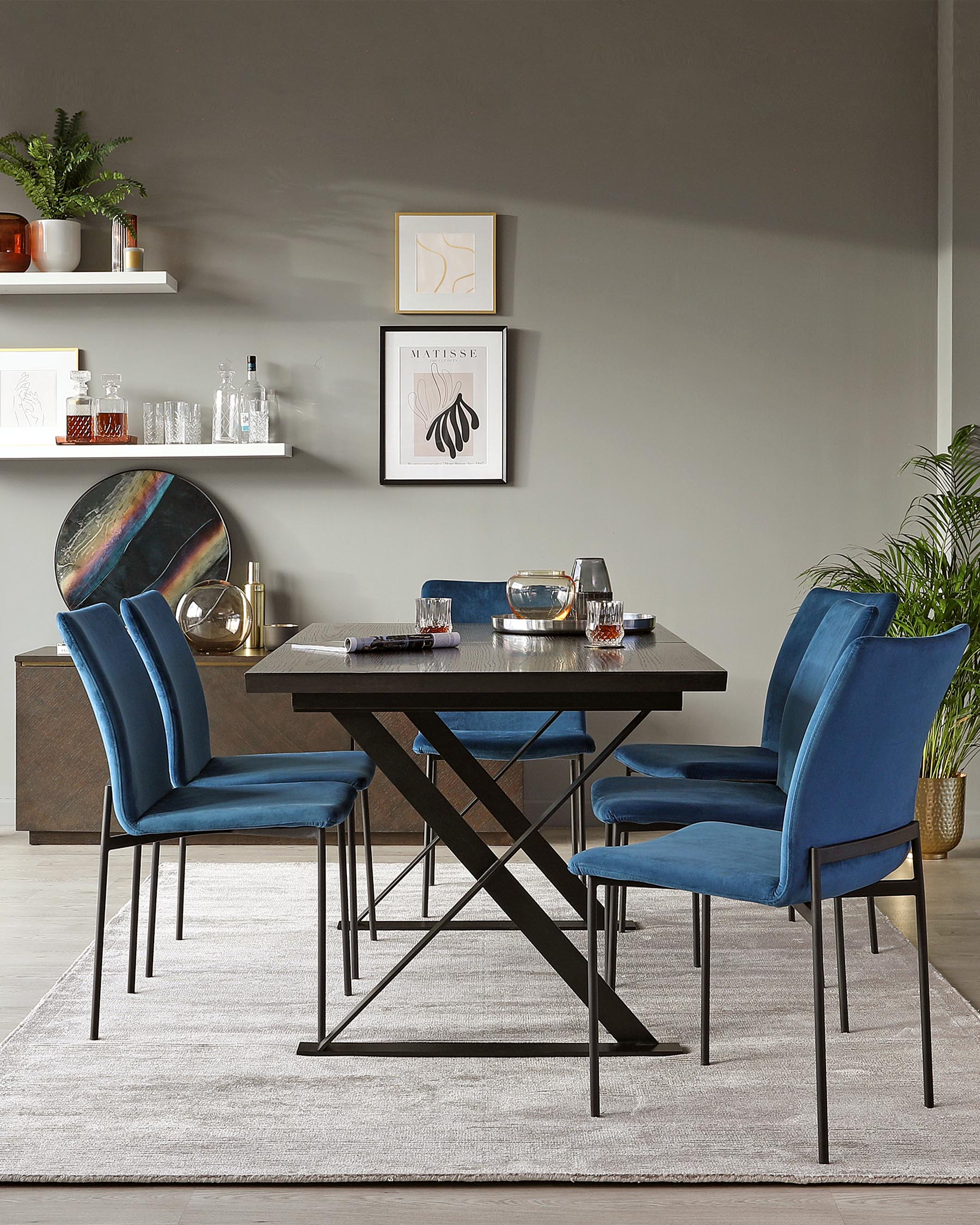 Contemporary dining room furniture featuring a rectangular wooden table with an X-shaped metallic black base. Accompanied by four plush velvet-upholstered dining chairs in rich blue with sleek black metal legs. The set is positioned on a light grey textured area rug, against a backdrop of elegant shelving and modern decor.