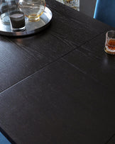 A modern dark wood dining table with a textured finish displaying a metal tray with a glass decanter, a black candle, and a glass cup, alongside a separate whiskey glass.