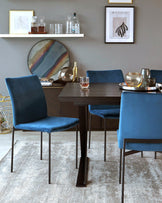 Minimalist dark wooden dining table with sleek square legs, accompanied by four plush blue velvet-upholstered chairs with slender metal legs. A textured light grey rug underlays the furniture set.