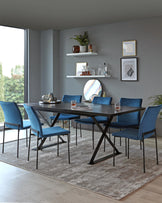 A modern dining room featuring a rectangular dark wood table with an intersecting black metal leg design, complemented by six plush blue upholstered dining chairs with black metal legs. On the wall are two white floating shelves with decorative items and framed artwork above. The arrangement is set on a textured grey area rug.