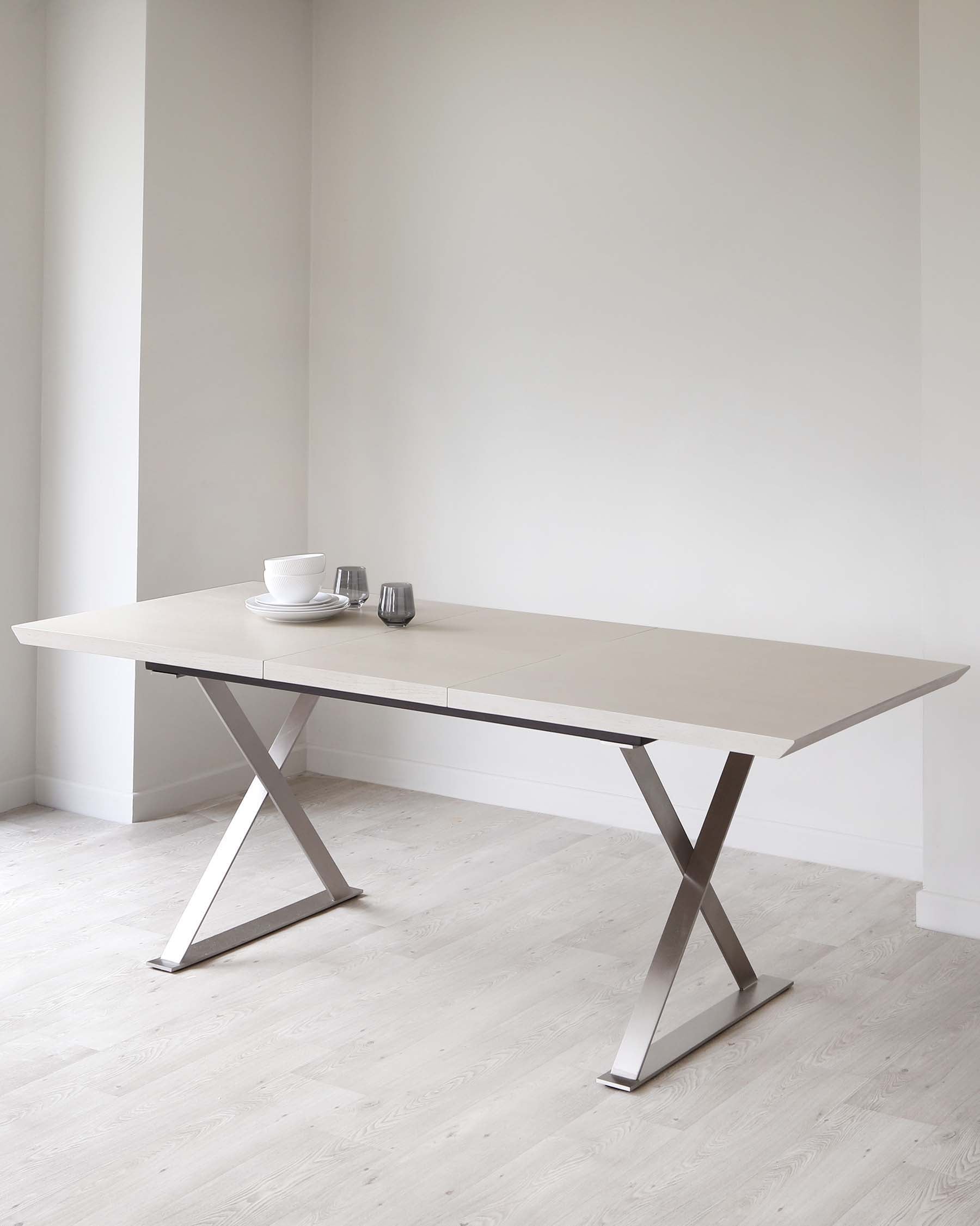 Modern dining table with a sleek, light wood top and minimalist silver X-shaped metal legs, set with dishes and glasses.