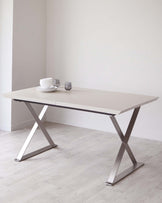 Modern dining table with a light wood surface and sleek, stainless steel X-frame legs, set with white dishware and glasses.