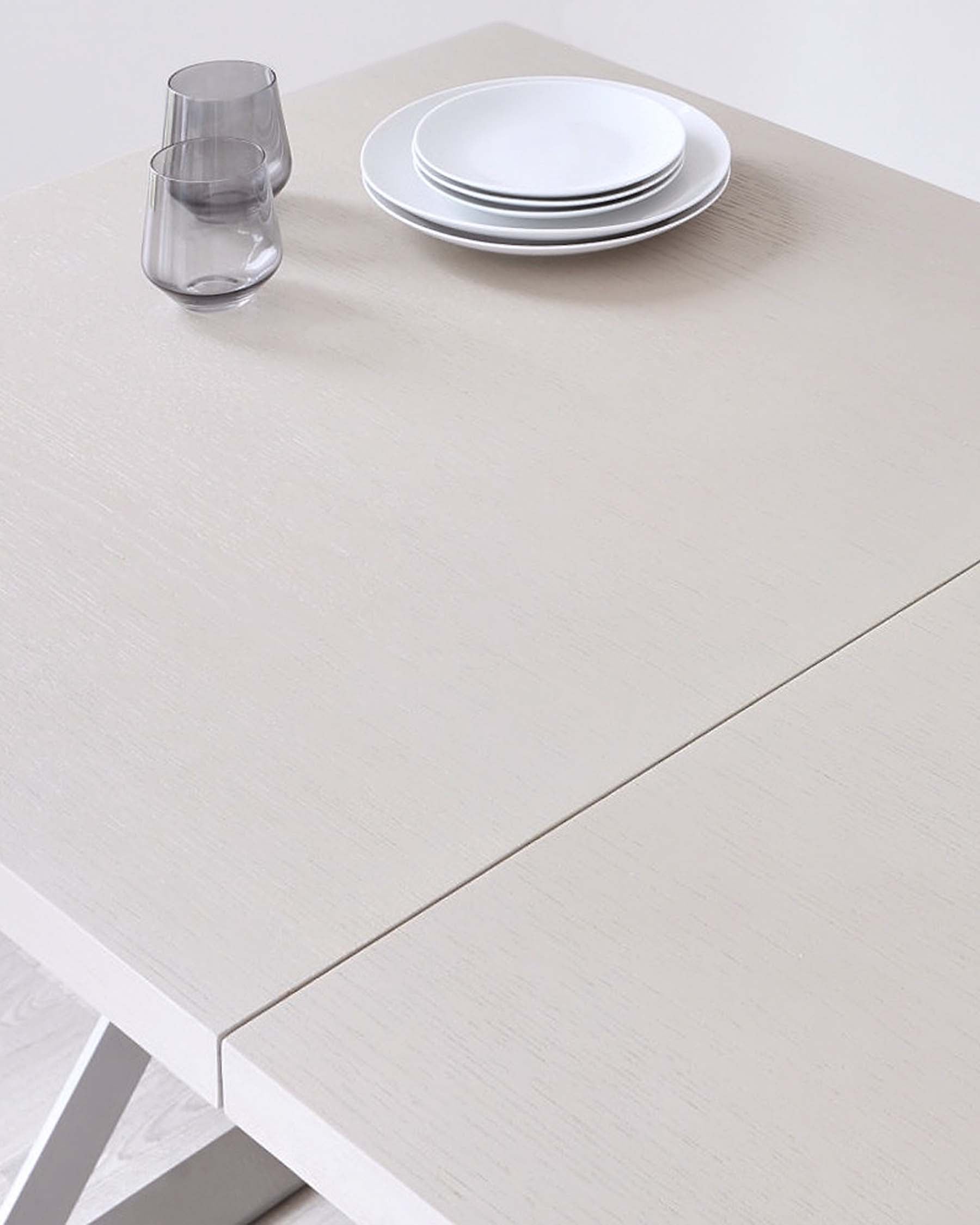 A light wood dining table features stacked white plates and two translucent glasses, creating a minimalist and modern setting.