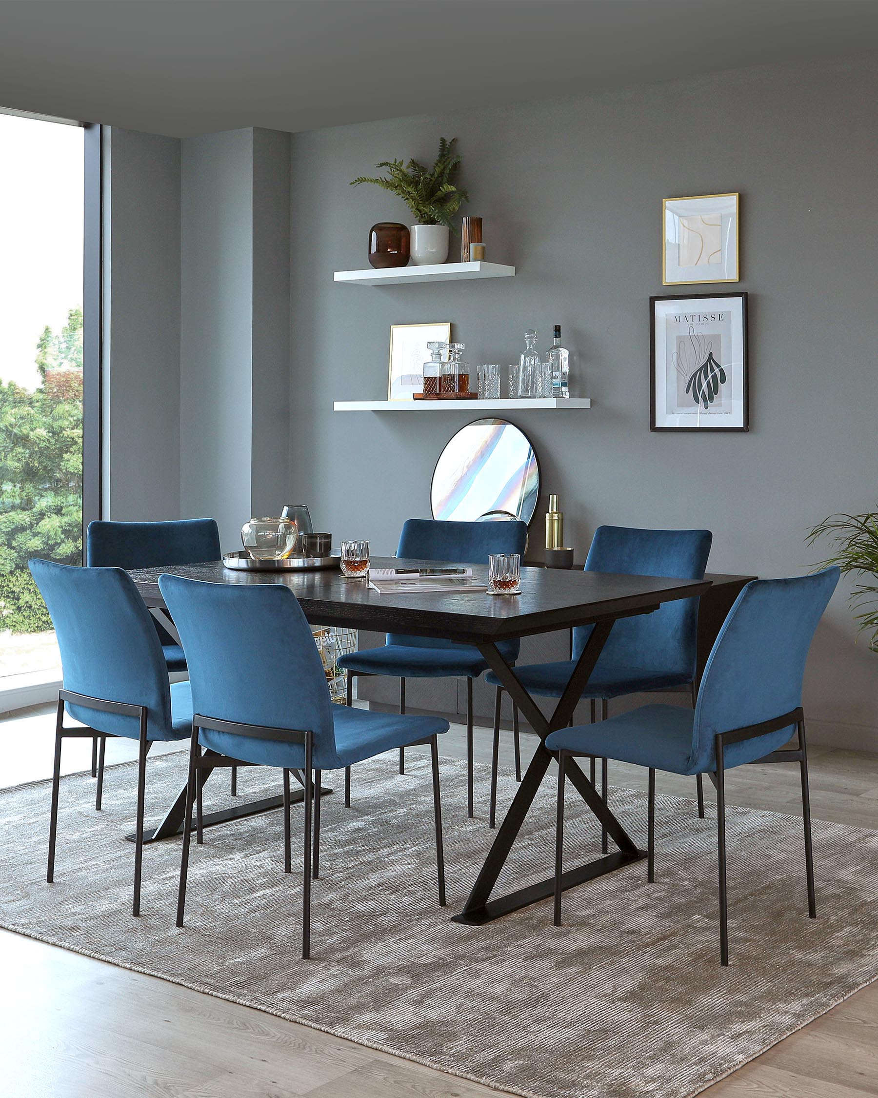 Modern dining room featuring a rectangular dark wood table with an X-shaped base, surrounded by six blue upholstered chairs with sleek black metal legs, all positioned on a textured grey area rug. Two white floating shelves adorn the wall above the table, displaying assorted decorative items.