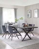 A modern dining room with a rectangular dark wood table that has a unique black X-shaped leg design on each end. Accompanying the table are four plush upholstered chairs with vertical stitching on the backrest, in a complementary shade of grey, each resting on black metal legs. A minimalist black console table is visible in the background, along with a simple, light grey area rug under the dining set.