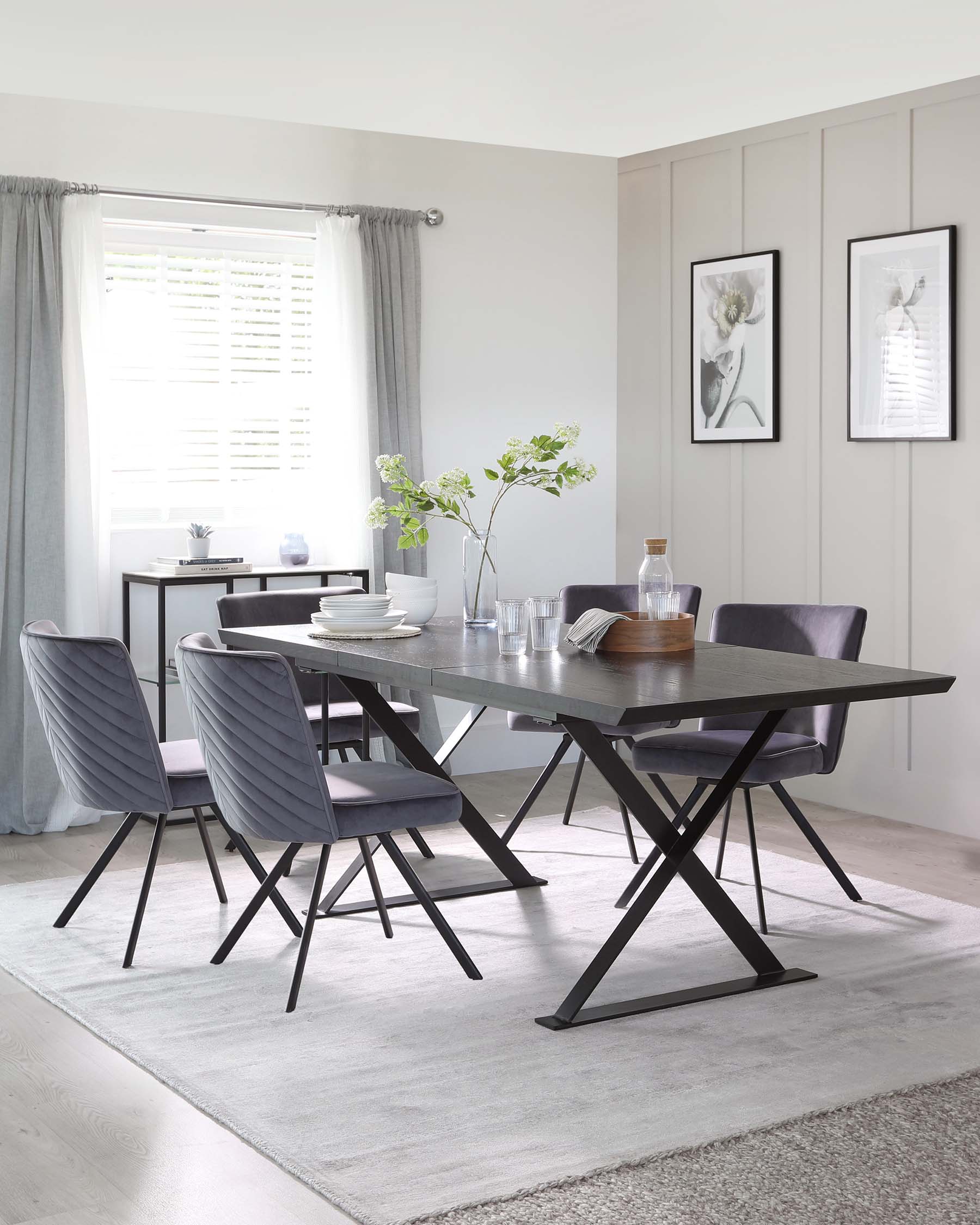 A modern dining room with a rectangular dark wood table that has a unique black X-shaped leg design on each end. Accompanying the table are four plush upholstered chairs with vertical stitching on the backrest, in a complementary shade of grey, each resting on black metal legs. A minimalist black console table is visible in the background, along with a simple, light grey area rug under the dining set.