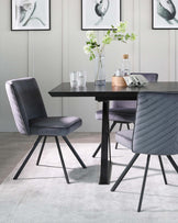 A modern dining set featuring a matte black rectangular table with solid angled legs, paired with two plush upholstered chairs in a deep grey tone. The chairs have a unique, vertical channel-tufted design on the backrest and are supported by black metal legs that match the table. The simple yet elegant setup is complemented by a neutral-toned area rug beneath.