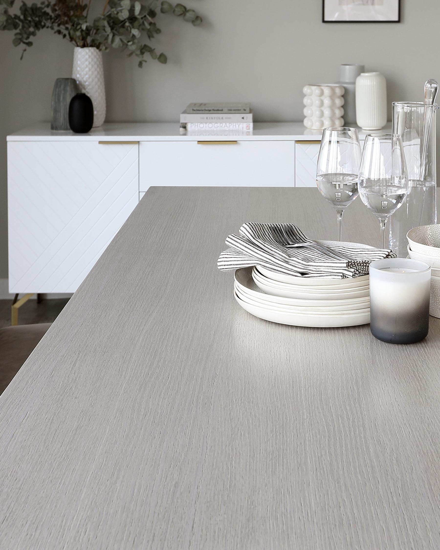 A modern dining table with a textured, light grey wood-grain finish, complemented by a sleek white sideboard featuring a geometric pattern on its doors. The sideboard is accessorized with a collection of decorative vases and books.