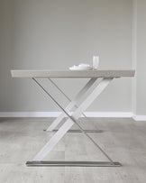 Modern minimalist dining table with a light wood grain finish and striking white crisscross metal legs, set against a neutral interior backdrop.