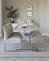 Modern minimalist dining room setup with a light wood rectangular table featuring a white geometric pattern on top and striking X-shaped metallic legs, complemented by two upholstered chairs with a luxurious velvet finish and sleek metal frames. The scene is anchored by a subtle textured area rug.