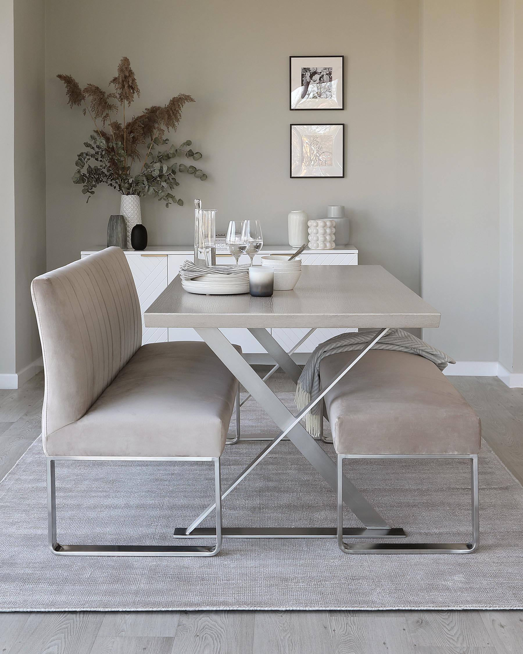 Modern minimalist dining room setup with a light wood rectangular table featuring a white geometric pattern on top and striking X-shaped metallic legs, complemented by two upholstered chairs with a luxurious velvet finish and sleek metal frames. The scene is anchored by a subtle textured area rug.