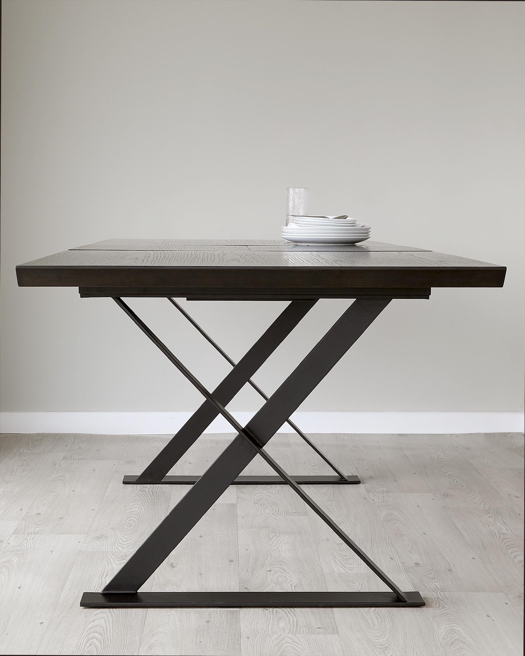 Modern dark wood dining table with a rectangular top and a unique crossed metal base, paired with simple white plates and a clear water glass, set against a neutral backdrop and light wooden flooring.