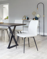 Contemporary dining room set featuring a minimalistic dark wooden table with a unique X-shaped leg design in black, complemented by a set of sleek white upholstered chairs with black metal legs, situated on a light wood laminate floor with a stylish black floor lamp arching over the scene.