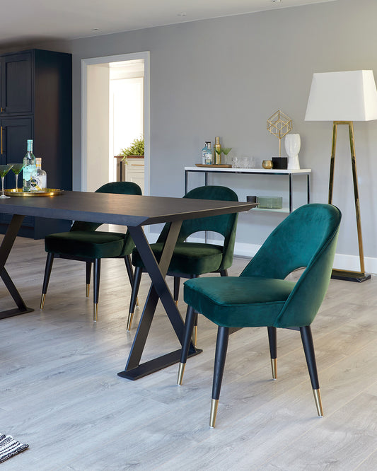 Contemporary dining room featuring a dark wooden table with an X-shaped base, accompanied by four luxurious dark green velvet chairs with black legs tipped with brass. A sleek sideboard with a glass shelf behind the dining set displays decorative items, complemented by a tall floor lamp with a white shade and brass stand in the background.