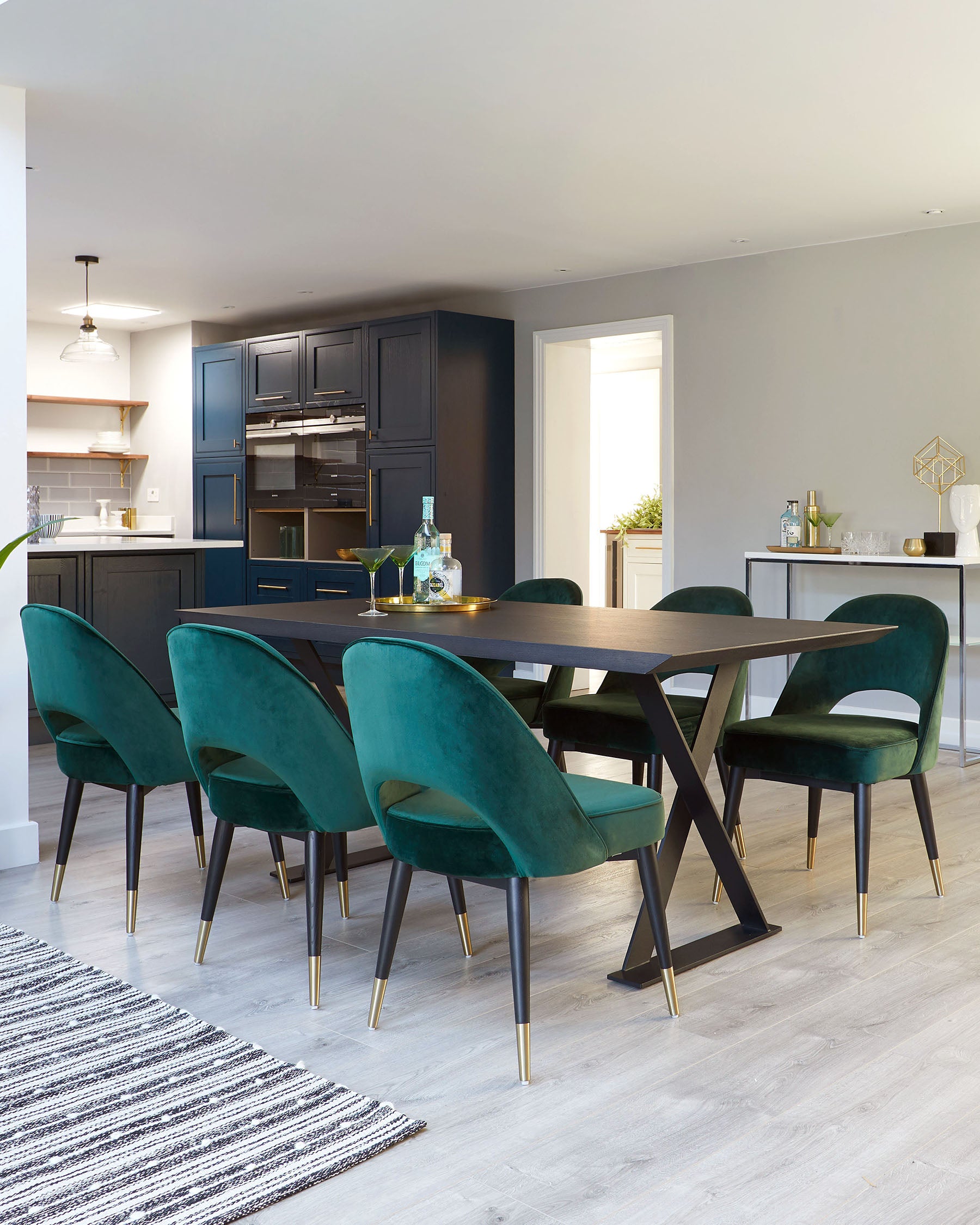 Elegant contemporary dining set featuring a rectangular dark wood table with a unique V-shaped leg design in a contrasting black and brass finish. Six plush velvet dining chairs in a rich forest green colour, with slim tapered legs tipped with brass accents, complement the table. The set offers a sophisticated, luxurious addition to any modern dining space.