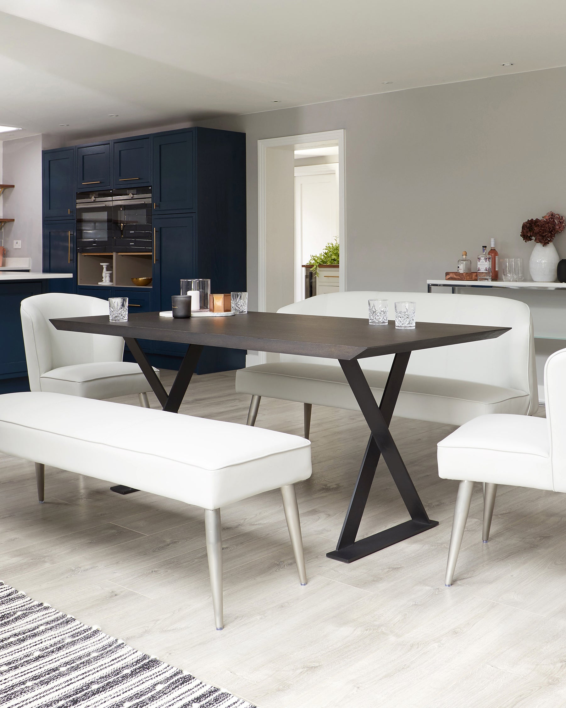 Modern dining room with a rectangular dark wood table featuring an X-shaped base, flanked by a white upholstered bench on one side and two white upholstered chairs with conical legs on the other.