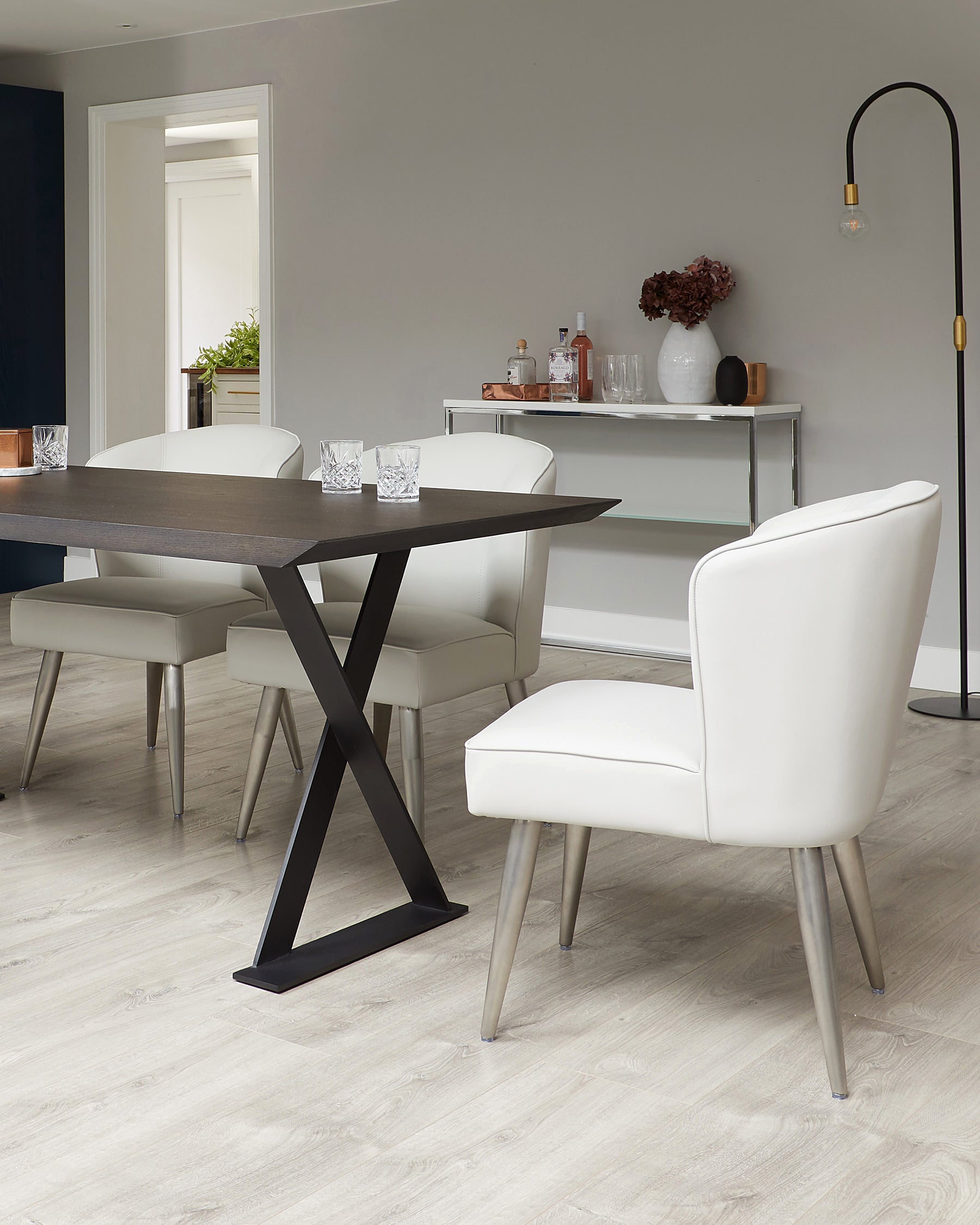 Modern dining room showcasing an elegant rectangular dark wood table with a unique black X-shaped base. Accompanied by four sleek, upholstered chairs in a white leather finish with metallic tapered legs. A minimalist sideboard with a glass shelf and metallic frame is placed against the wall, decorated with a vase, bottles, and glasses, complementing the contemporary aesthetic of the set-up. A tall, curved floor lamp with a gold accent stands nearby, adding warmth to the scene.