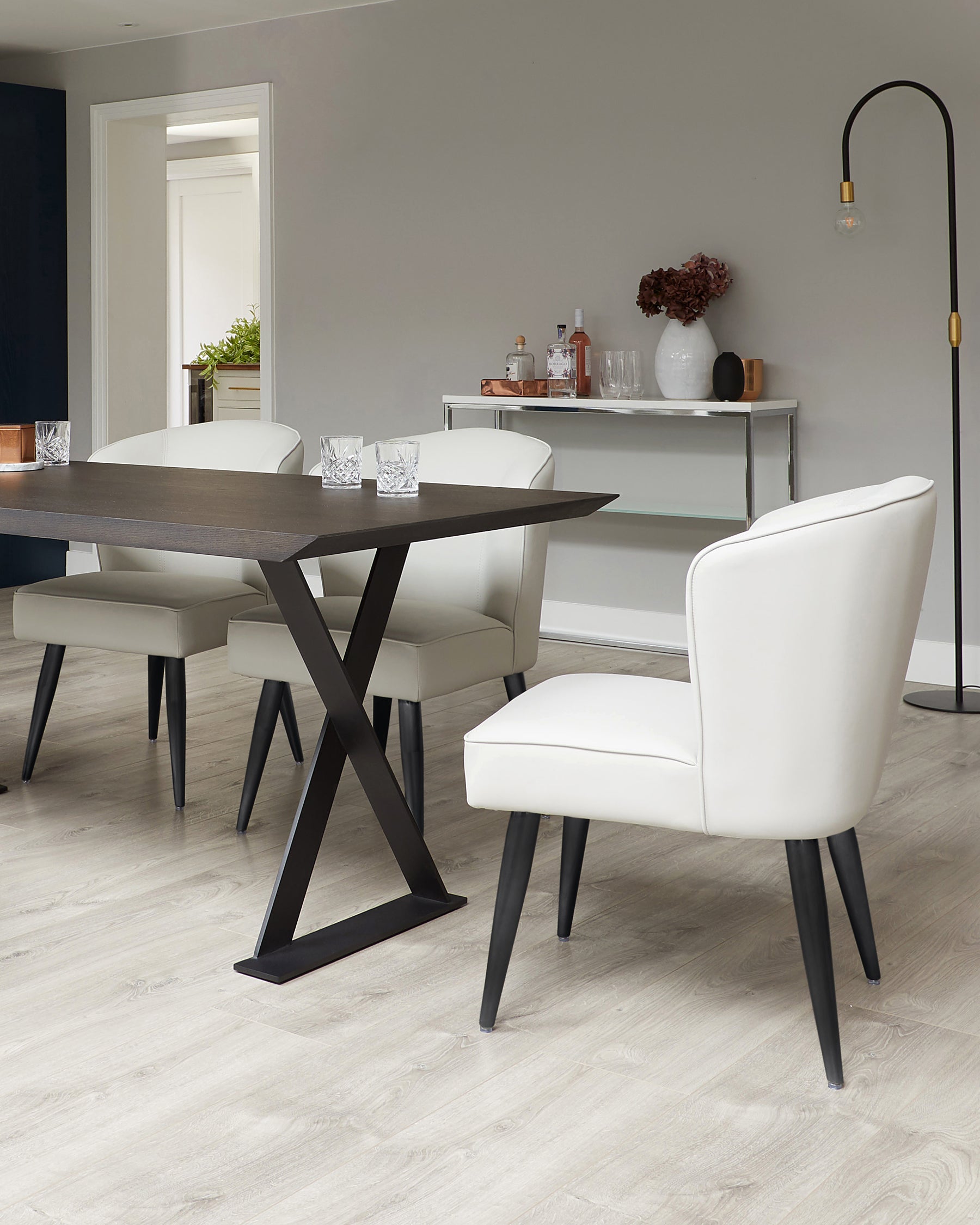 Modern dining room setup featuring a dark wooden table with an X-shaped metal base, surrounded by four cream upholstered chairs with black tapered legs. A minimalist white console table with glass shelves is in the background, adorned with decorative bottles, vases, and a bouquet. A sleek, arched floor lamp with a gold accent complements the scene.