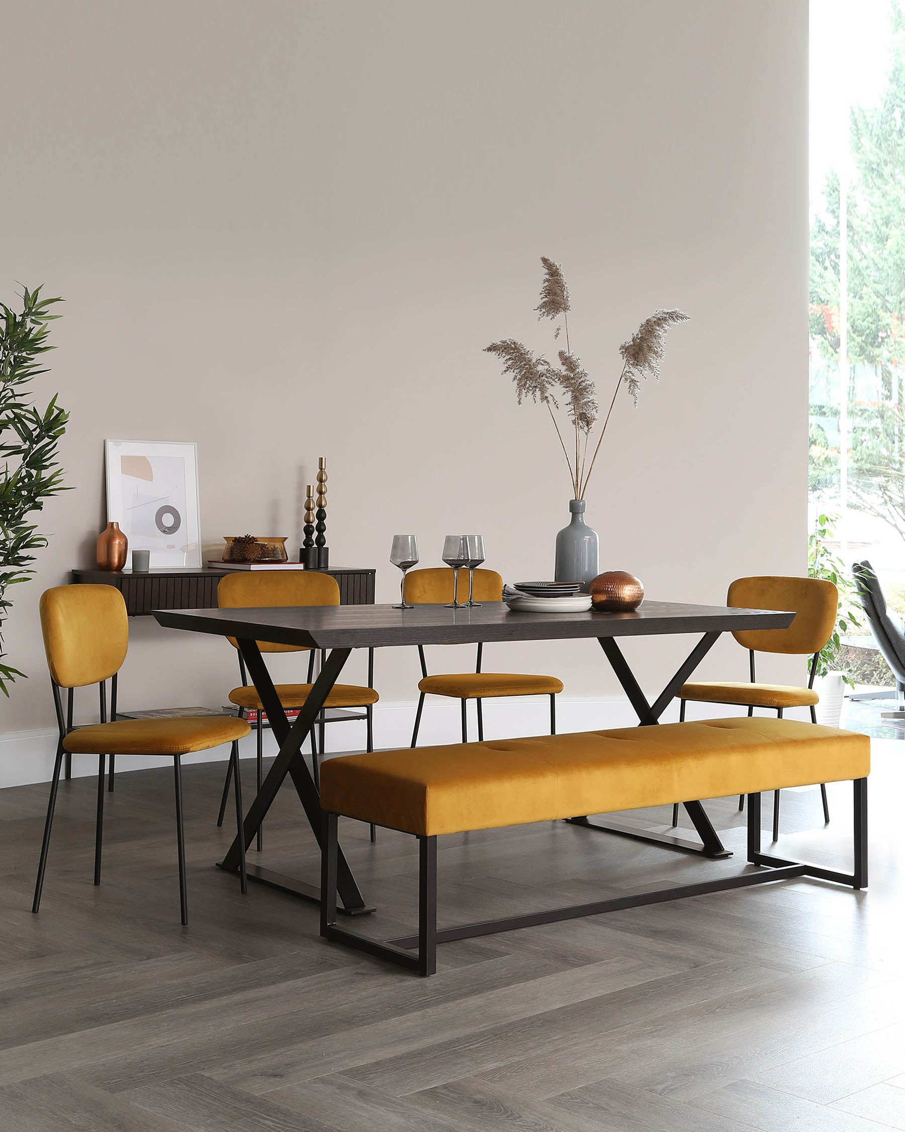 Modern dining room set featuring a rectangular dark wood table with black metal legs in an X-shape configuration. The set includes four mustard yellow upholstered chairs with slim black metal legs and a matching mustard yellow upholstered bench with a black metal frame. The table is partially set with plates, glasses, and a decorative grey vase with pampas grass, complemented by accents on a side console in the background.