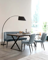 Modern dining room furniture featuring a rectangular wooden table with a dark finish and unique V-shaped metal legs, complemented by a coordinating bench and two padded chairs with a sophisticated vertically channelled teal upholstery. An elegant black floor lamp with a large curved neck and an oversized shade towers over the setting, completing the contemporary aesthetic.