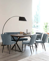 Modern dining room with a dark wood table featuring a unique X-shaped base, surrounded by four plush upholstered dining chairs in a soft teal fabric with vertical stitching on the backrest. Overhead is an elegant, black, arc floor lamp with a large shade, adding a contemporary touch to the space.