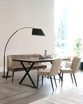 Modern dining area featuring a dark wood table with X-shaped black metal legs, surrounded by four upholstered beige dining chairs with vertical stitching and slender black legs. An oversized black floor lamp with an arching design adds a dramatic touch over the scene.