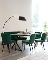 Modern dining room featuring a dark wooden rectangular table with a unique X-shaped metal base, surrounded by four plush, channel-tufted chairs in a vibrant emerald green velvet fabric with slim black legs. An elegant, oversized black arc floor lamp with a large dome shade creates a focal point above the setup.