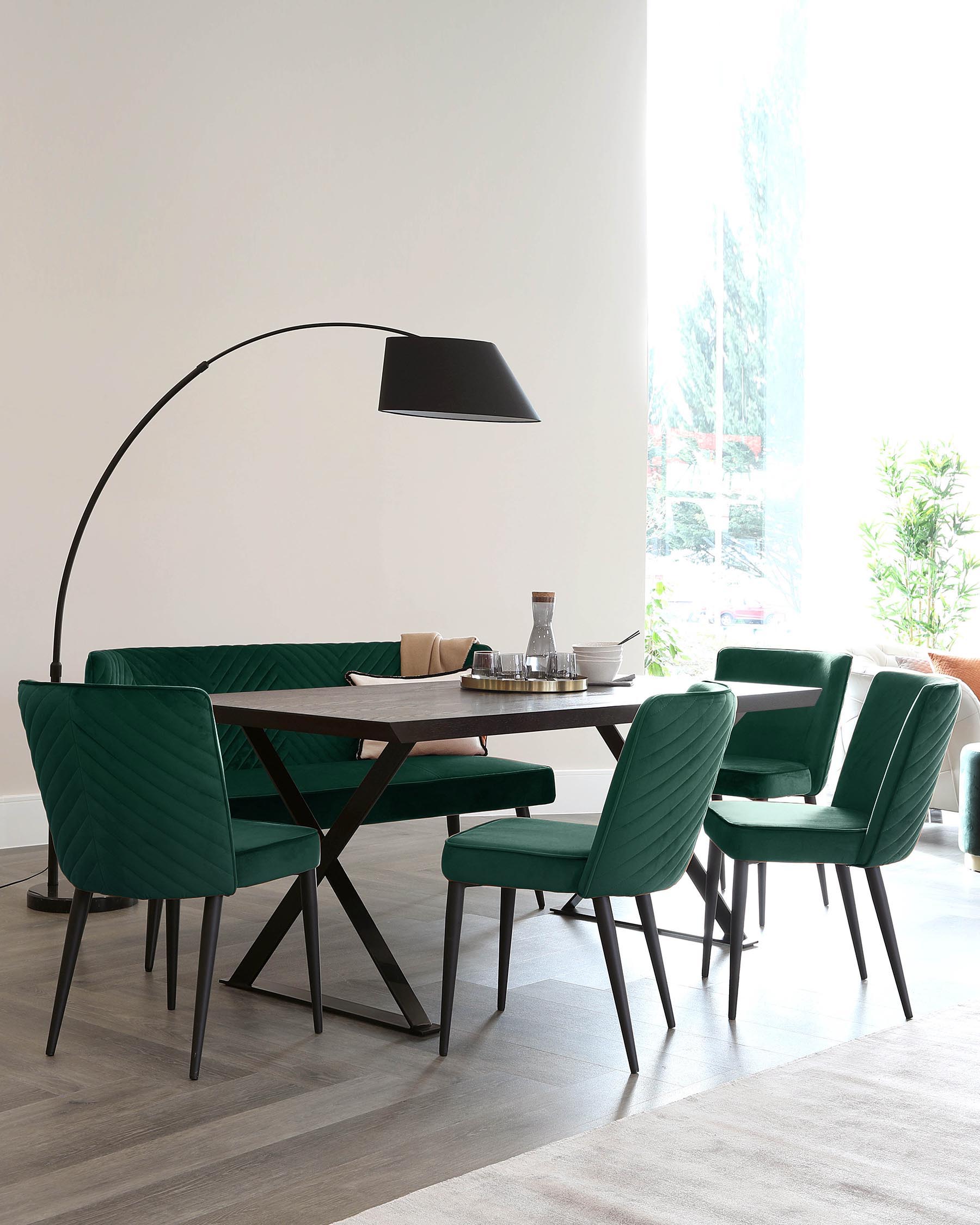 Modern dining room featuring a dark wooden rectangular table with a unique X-shaped metal base, surrounded by four plush, channel-tufted chairs in a vibrant emerald green velvet fabric with slim black legs. An elegant, oversized black arc floor lamp with a large dome shade creates a focal point above the setup.