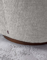 Close-up view of a piece of furniture featuring a textured light-coloured fabric upholstery with a visible brand label, and a rounded wooden base on a textured light carpet.