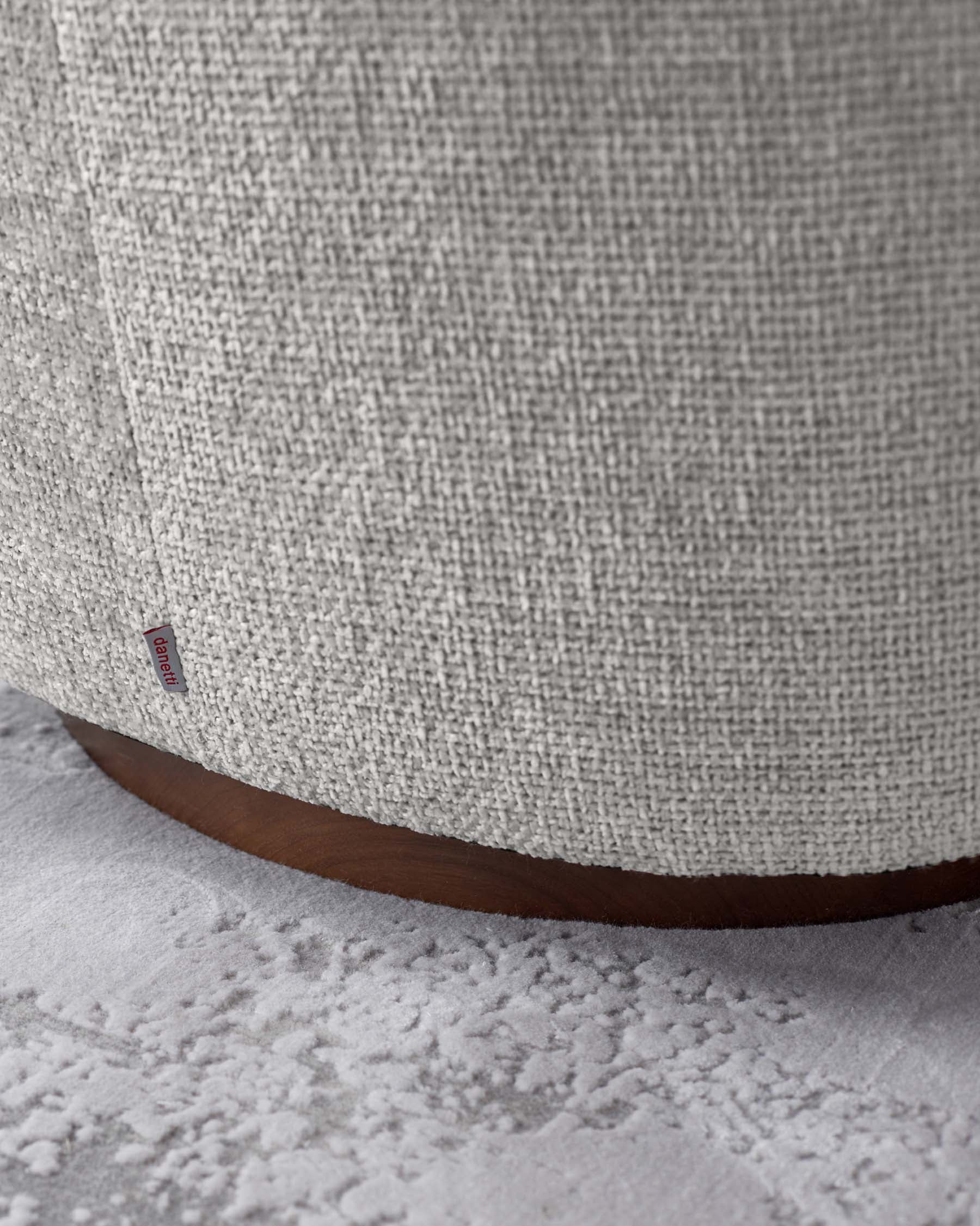 Close-up view of a piece of furniture featuring a textured light-coloured fabric upholstery with a visible brand label, and a rounded wooden base on a textured light carpet.