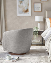 Contemporary grey fabric swivel chair with a rounded backrest, visible black zipper detailing, and a dark wood circular base, accompanied by a modern white table lamp on a glass-top side table with metallic accents. A draped throw blanket and decorative pillows are partially visible, suggesting a comfortable and stylish living space.