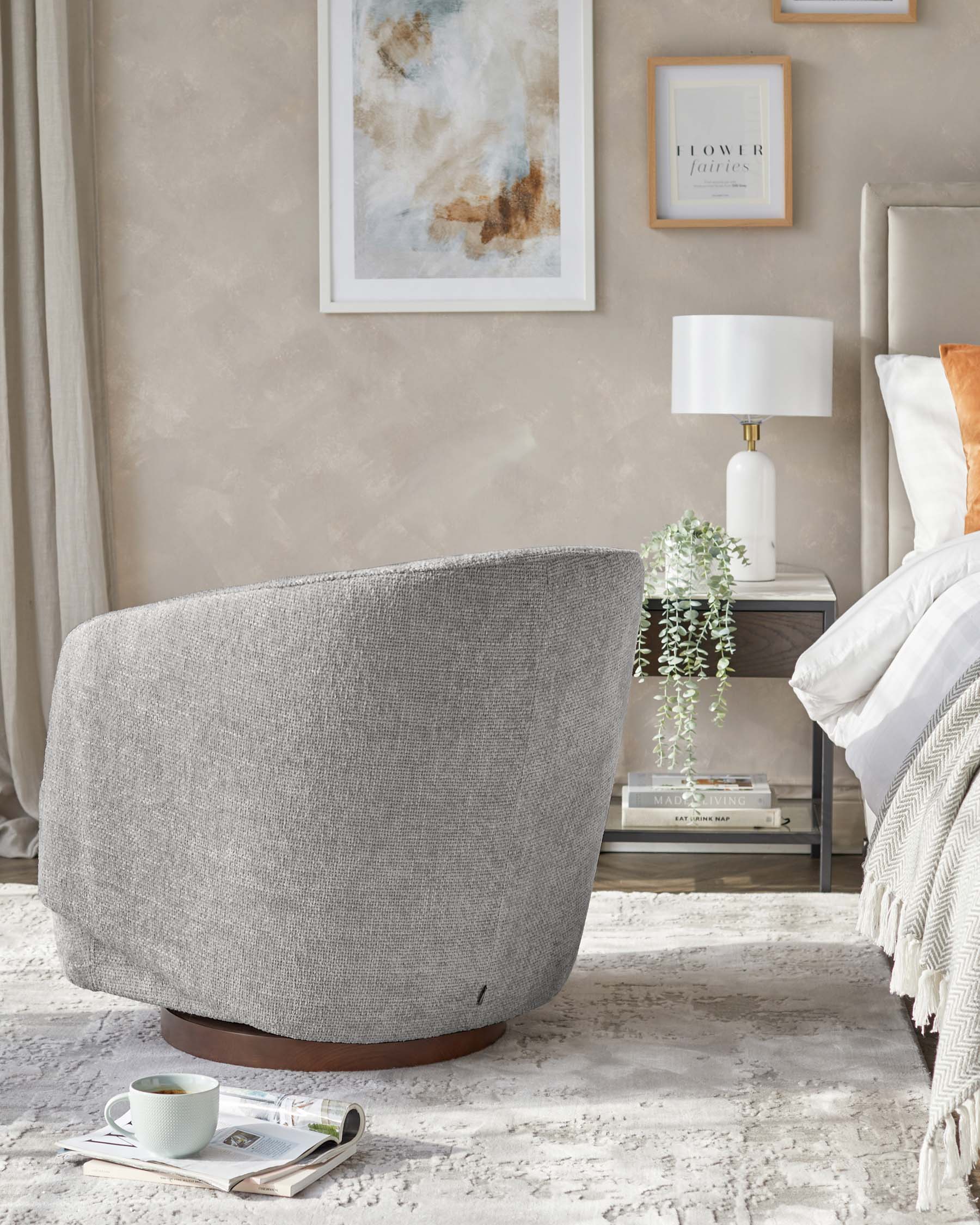 Contemporary grey fabric swivel chair with a rounded backrest, visible black zipper detailing, and a dark wood circular base, accompanied by a modern white table lamp on a glass-top side table with metallic accents. A draped throw blanket and decorative pillows are partially visible, suggesting a comfortable and stylish living space.