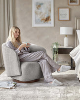 A contemporary styled round swivel armchair upholstered in textured light grey fabric, featuring a deep seat and flared armrests on a contrasting dark wood circular base. Alongside is a minimalist dark wood bedside table with a single drawer, displaying a modern white cylindrical table lamp, topped by a simple shade and a potted green plant, against a neutral-coloured bedroom setting.