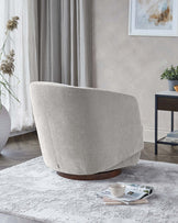 A contemporary light grey fabric upholstered armchair with a curved backrest and a unique circular wooden base, positioned on a textured white area rug with a small wooden side table to the right bearing a potted plant. In the foreground, there is a casually placed set of magazines and an open book with a ceramic cup on top.