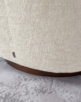 Close-up view of the lower section of a beige upholstered piece with a textured fabric finish and a curved dark wooden base, set against a grey carpeted floor with a subtle pattern. A small red label with text is visible on the fabric.