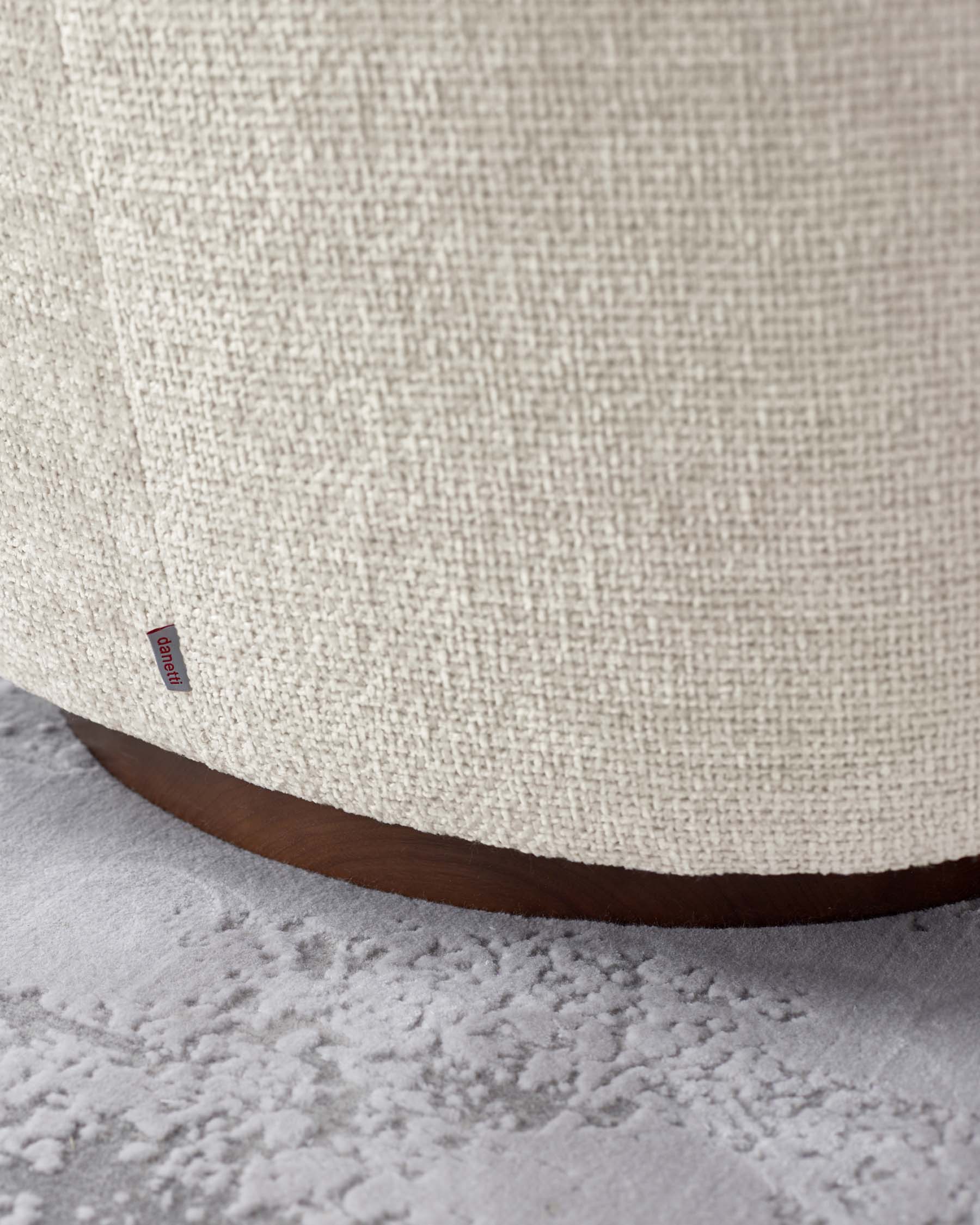 Close-up view of the lower section of a beige upholstered piece with a textured fabric finish and a curved dark wooden base, set against a grey carpeted floor with a subtle pattern. A small red label with text is visible on the fabric.