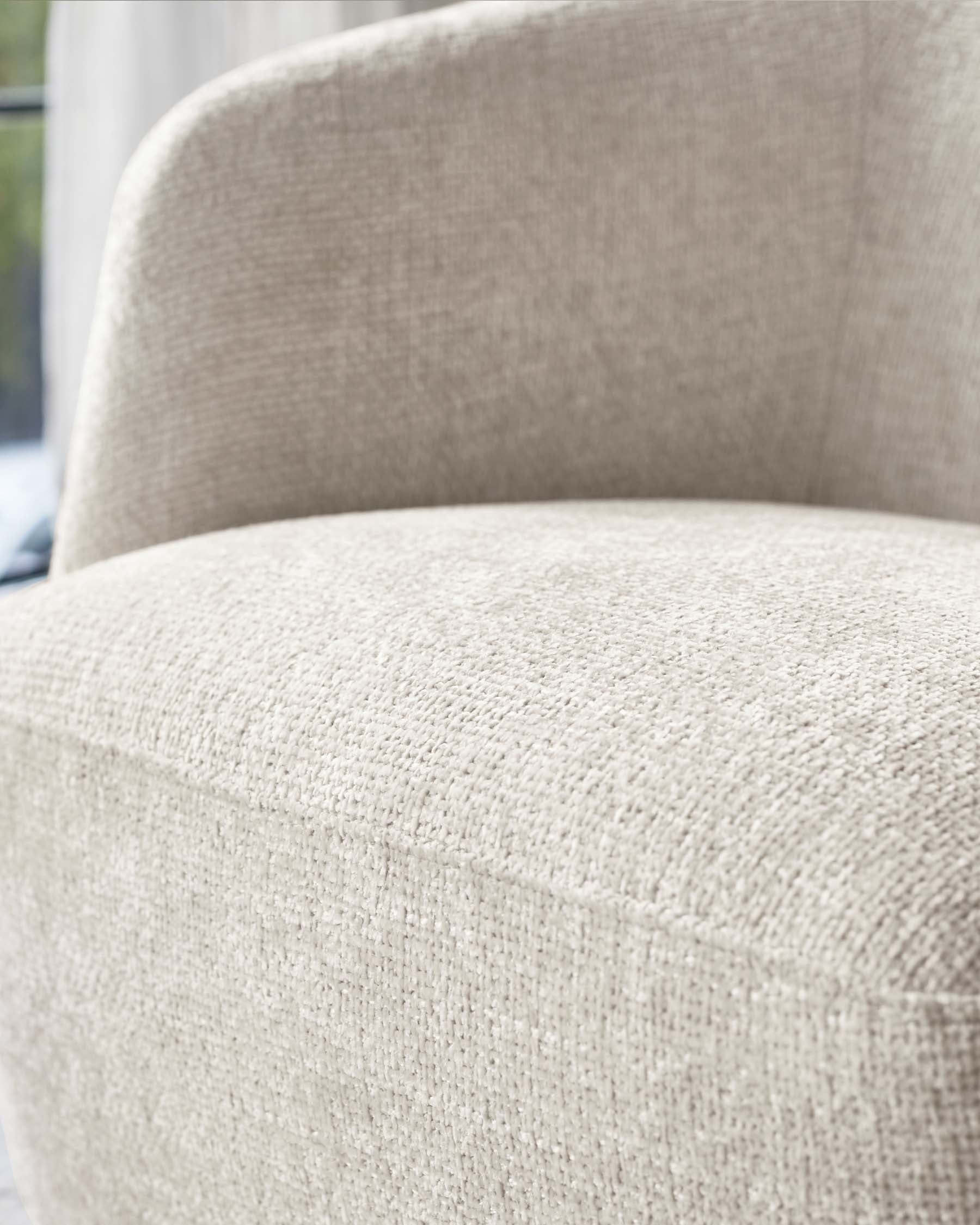 Close-up of a beige textured fabric armchair with a focus on the upholstery texture and armrest. The chair is partially visible suggesting a contemporary design with a soft, inviting appearance, set against a light-filled room with sheer curtains in the background.