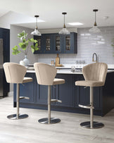 Modern kitchen with three beige upholstered bar stools featuring curved backrests and footrests, set against a kitchen island with dark blue cabinetry and white countertop.