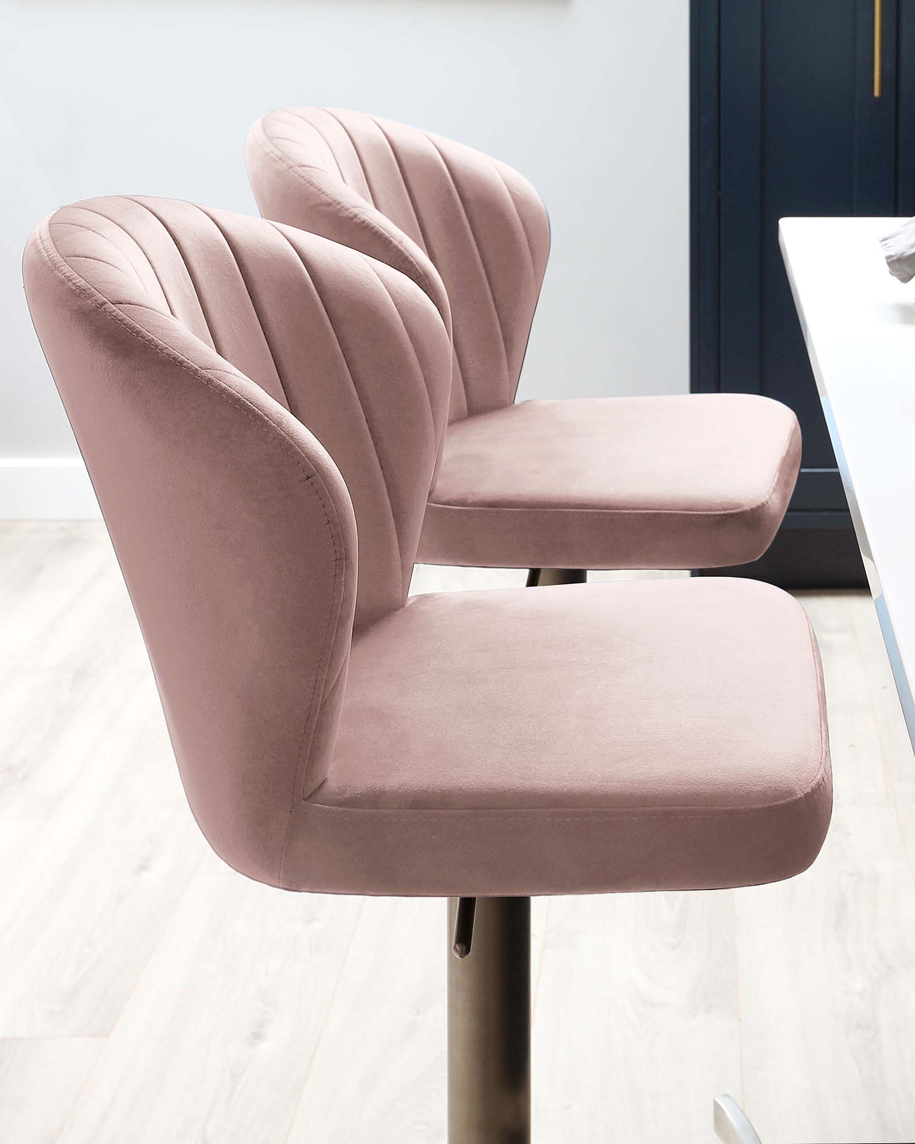 Elegant blush pink velvet bar stools with channel tufted backrests and comfortable cushioned seats, mounted on sleek, adjustable metal bases.